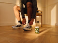 A person sitting on a wooden chair in a warm, sunlit room, tying the laces of white and black Adidas running shoes. A clear water bottle with a green and white Adidas logo sits on the wooden floor nearby.