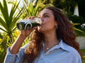 A woman with long wavy brown hair drinks from a clear plastic bottle with a white logo, set against a background of green plants and outdoor foliage.