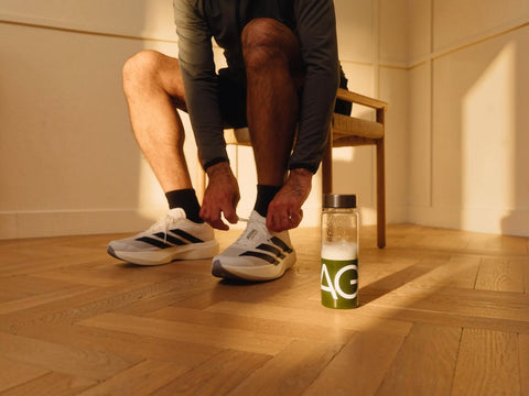 A person sitting on a wooden chair in a warm, sunlit room, tying the laces of white and black Adidas running shoes. A clear water bottle with a green and white Adidas logo sits on the wooden floor nearby.