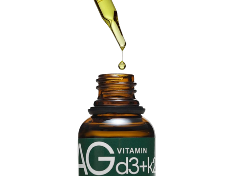 An amber glass dropper bottle with a green label reading 'AG VITAMIN d3+k2' on a white background, with a glass dropper above dispensing a drop of golden oil.