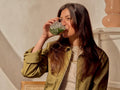 A woman with long dark hair wearing an olive green jacket and a cream top drinks from a textured clear glass cup with green leaves inside, set against a warm beige background.