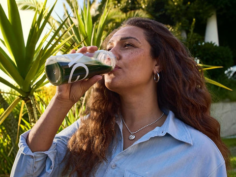 A woman with long wavy brown hair drinks from a clear plastic bottle with a white logo, set against a background of green plants and outdoor foliage.
