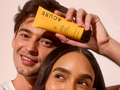Person holding a yellow Acure product above another person's head against a plain background