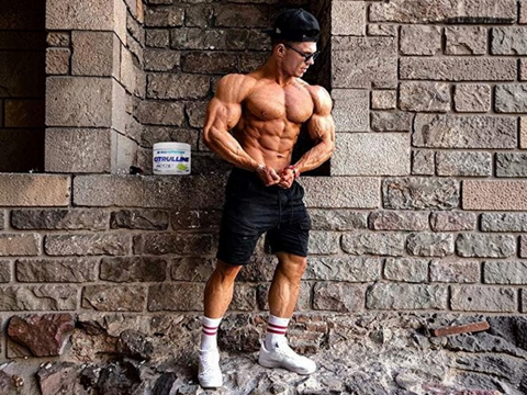 Muscular man posing in front of a brick wall with a supplement container on a ledge.