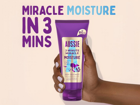 Aussie 3 Minute Miracle Moisture Deep Treatment tube held by a hand with text 'Miracle Moisture in 3 Mins' on a beige background.