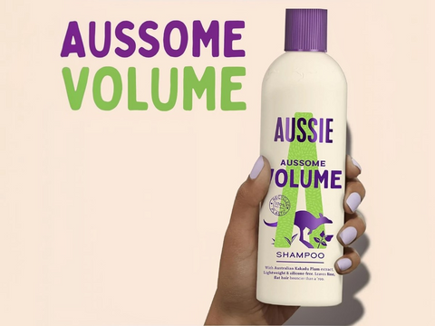 Aussie Aussome Volume shampoo bottle held by a hand with text on a beige background
