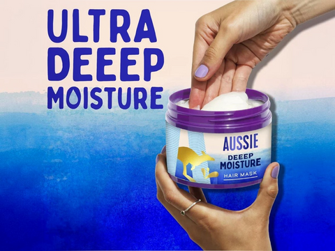 Aussie Deep Moisture hair mask jar with hands holding it against a blue and pink gradient background.