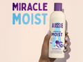 Aussie Miracle Moist shampoo bottle held by a hand with a pink background and text.