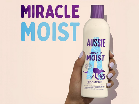 Aussie Miracle Moist shampoo bottle held by a hand with a pink background and text.