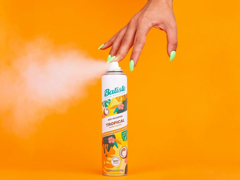 Hand holding a can of Batiste Tropical hair spray against an orange background