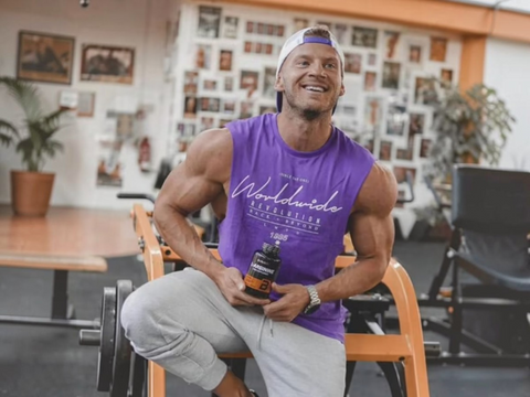 Man in purple sleeveless shirt holding a supplement bottle in a room with plants and furniture.