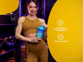 Woman holding a supplement shaker in a gym setting with product information on a yellow background