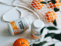 Two bottles of vitamin C supplements on a surface with oranges and a checkered cloth in the background.