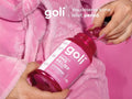 Person holding a pink bottle of goli PMS Relief supplement with a pink robe in the background.