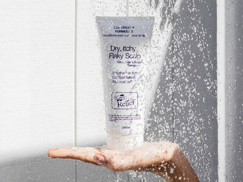 Hand holding a bottle of 'Dry, Itchy Flaky Scalp' treatment under running water.