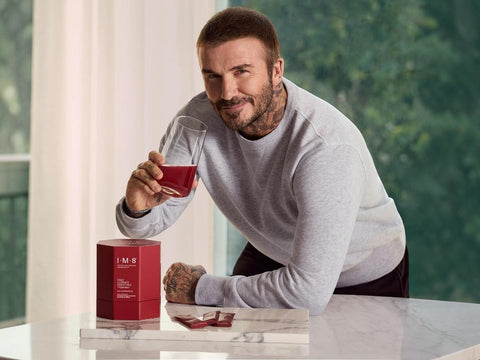 A man in a light gray sweatshirt leans over a white marble table, holding a glass of deep red liquid. A red rectangular product box with white text sits on the table beside him, with a few small red packets in front of it.