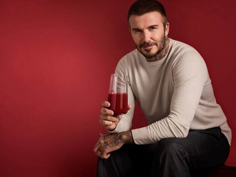 A man with tattoos wearing a beige long-sleeve shirt and dark pants sits against a solid red background, holding a clear glass filled with red juice.