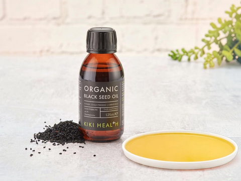 Bottle of Kiki Health Organic Black Seed Oil on a light surface with black seeds and a yellow substance.
