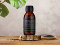 Bottle of KIKI Health Organic Black Seed Oil on a wooden surface with black seeds and green leaves.
