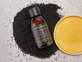 Bottle of KIKI Health Organic Black Seed Oil surrounded by black seeds on a light surface.