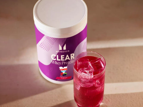 Glass of clear, purple Myprotein Vimto protein drink next to the powder tub.