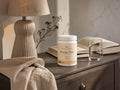 A white tub of Neuroline Chamomile 'Boxine Pure' herbal supplement sits on a dark wooden nightstand in a cozy, warmly lit bedroom setting, surrounded by a lamp, an open book, a glass of water, and a beige linen throw.