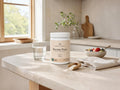 A white tub of Nordic Omega Bovine Pure supplement powder sits on a light stone kitchen counter beside a glass of water, a small bowl of fruit, and a cloth scoop of powder. The scene is set in a bright, modern kitchen with natural light.