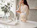 A woman in a cream robe stands in a bright bathroom or spa-like interior, holding a small container near her face. In the foreground on a white marble counter is a white tub labeled 'Nordic Omega Bovine Pure' with a folded white towel, a small round mirror, a glass of water, and a vase with eucalyptus branches.