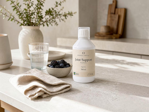 A bottle of Pure Encapsulations Joint Support liquid supplement placed on a light stone countertop in a bright kitchen setting, surrounded by a glass of water, a bowl of blackberries, and a folded beige cloth.