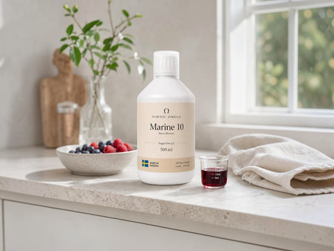 A bottle of Organic Omega Marine 10 liquid supplement sits on a light stone countertop in a bright kitchen setting, with a bowl of berries, a glass of dark liquid, and a folded towel nearby.