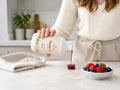 A person in a cream ribbed sweater and matching apron pours a dark red liquid from a white bottle labeled Marine 10 into a small cup on a kitchen counter, with a folded cloth and a bowl of berries nearby.