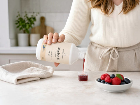 A person in a cream ribbed sweater and matching apron pours a dark red liquid from a white bottle labeled Marine 10 into a small cup on a kitchen counter, with a folded cloth and a bowl of berries nearby.