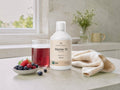 A bottle of Nordic Omega Marine 10 liquid supplement stands on a light stone countertop beside a glass of dark red liquid, a small bowl of berries, and a folded cream cloth. The background shows a softly blurred kitchen or bathroom setting with a window and vase.