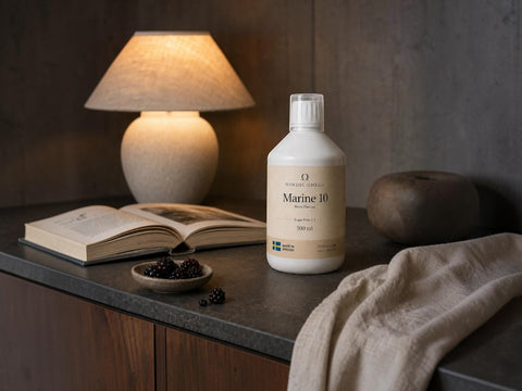 A bottle of Nordic Naturals Marine 10 seawater conditioner stands on a dark countertop in a styled home setting, alongside an open book, a lit table lamp, a small dish of blackberries, and a beige cloth.