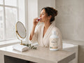 A woman in a cream robe stands in a bright bathroom, applying a pinkish-purple cosmetic or skincare product from a small glass tube. On the bathroom counter beside her is a white bottle labeled 'Marine 10' and a round mirror.