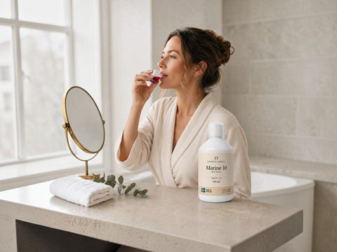 A woman in a cream robe stands in a bright bathroom, applying a pinkish-purple cosmetic or skincare product from a small glass tube. On the bathroom counter beside her is a white bottle labeled 'Marine 10' and a round mirror.