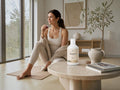 A woman in beige activewear sits on a yoga mat by a large window, holding a small glass. A white bottle labeled Marine 30 is placed on a round stone table in front of her, alongside a book and ceramic decor, in a bright, modern interior.