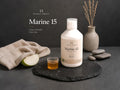 Nordic Omega Marine 15 liquid supplement bottle displayed on a dark slate tray with a folded beige towel, a small glass of amber liquid, a sliced green apple, and a ceramic vase with dried grass against a dark gray background.