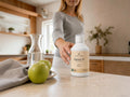 A woman in a gray long-sleeve top stands in a kitchen setting and reaches toward a white bottle labeled 'Nordic Omega Marine 15' placed on a countertop beside green apples and a glass carafe.