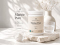 Nordic Omega Marine Pure collagen powder in a white jar, styled on a stone slab with a small glass bowl of powder, a ribbed glass, and a ceramic vase in a bright, sunlit setting.