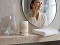 A woman in a white robe stands in front of a bathroom mirror, touching her face. On the countertop beside her is a jar labeled 'Omega Marine Pure' and a folded white towel, with a glass vase to the left.