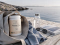 Nordic Omega Marine Pure supplement powder in a white jar, displayed inside a beige canvas tote bag on a wooden dock with a calm sea and rocky shoreline in the background. A glass water bottle, sunglasses, and a book are also visible in this lifestyle setting.