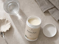 A jar of Nordic Omega Marine Pure supplement powder, shown open with its lid beside it on a light stone surface, with a glass of water, a spoon, and a folded cloth nearby.