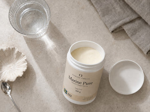 A jar of Nordic Omega Marine Pure supplement powder, shown open with its lid beside it on a light stone surface, with a glass of water, a spoon, and a folded cloth nearby.
