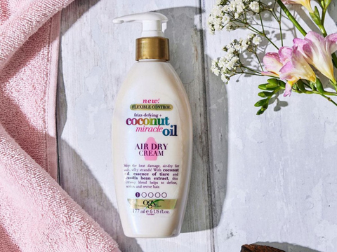 Bottle of OGX Coconut Miracle Oil Air Dry Cream with a pink towel and flowers on a wooden surface
