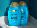 Two bottles of OGX shampoo and conditioner on a white surface with a blue background