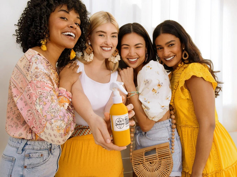 Four women posing together, holding a bottle of juice, with a neutral background.