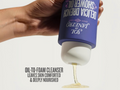 Hand holding a blue bottle of Delicia Drench shower oil with white foam being squeezed out.