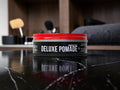 A tin of B10 B10 Deluxe Pomade with a bright red lid and black label, sitting on a glossy black marble surface with a blurred bathroom background.