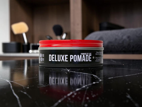 A tin of B10 B10 Deluxe Pomade with a bright red lid and black label, sitting on a glossy black marble surface with a blurred bathroom background.